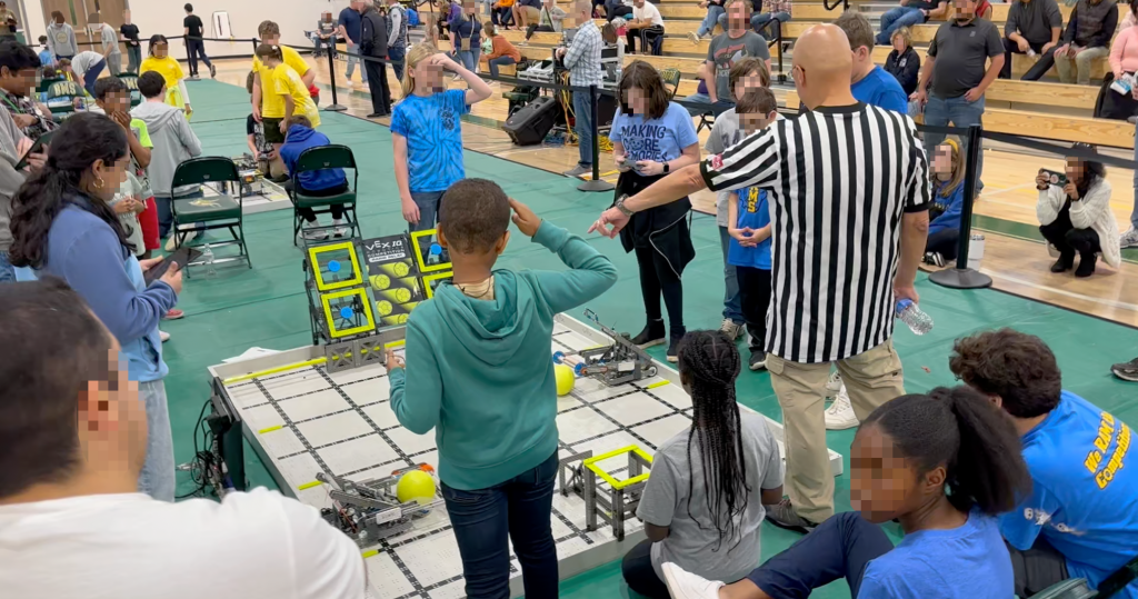 Children and adults gather around a robotics competition field with robots and colored balls, supervised by a referee.